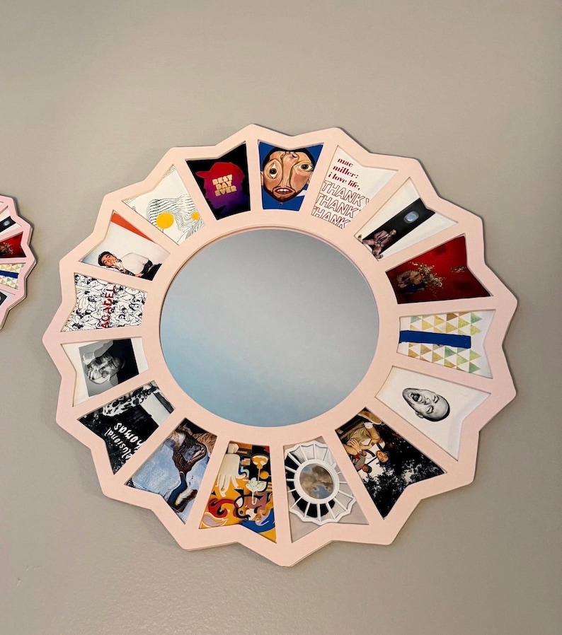 May include: A circular wall mirror with a light pink, star-shaped frame. The frame features sixteen individual compartments, each containing a different small square image or graphic print, surrounding a central circular reflective glass surface.