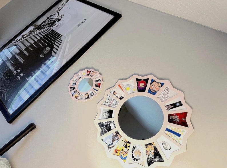 May include: Two circular mirrors with scalloped, light-coloured frames. The larger mirror features a border of small, colourful rectangular images arranged in a radial pattern. A smaller, matching mirror sits above it, also surrounded by tiny images.