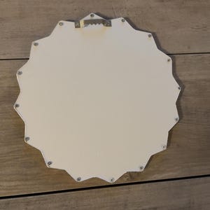 May include: A white, star-shaped wall plaque with a smooth, flat surface. The back features a metallic, serrated sawtooth hanger centred at the top for mounting. The edges of the star are defined by multiple points, and small silver fasteners are visible.