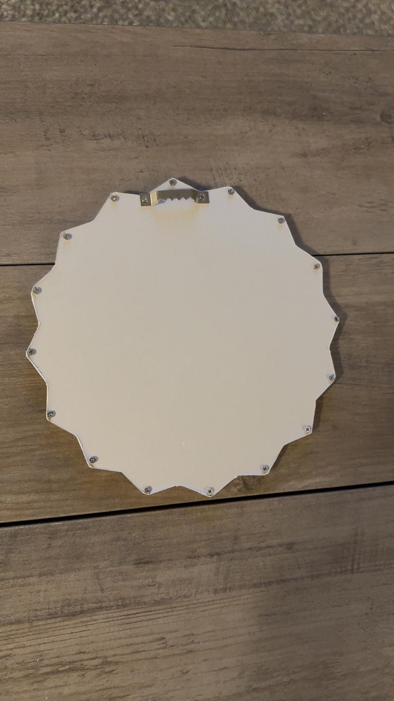 May include: A white, star-shaped wall plaque with a smooth, flat surface. The back features a metallic, serrated sawtooth hanger centred at the top for mounting. The edges of the star are defined by multiple points, and small silver fasteners are visible.