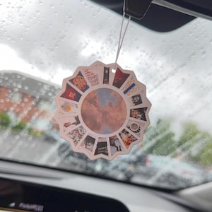 May include: A circular car air freshener featuring a scalloped edge and a central image of a cloudy blue sky. The border is composed of a collage of various small square album cover artworks arranged in a radial pattern around the centre.