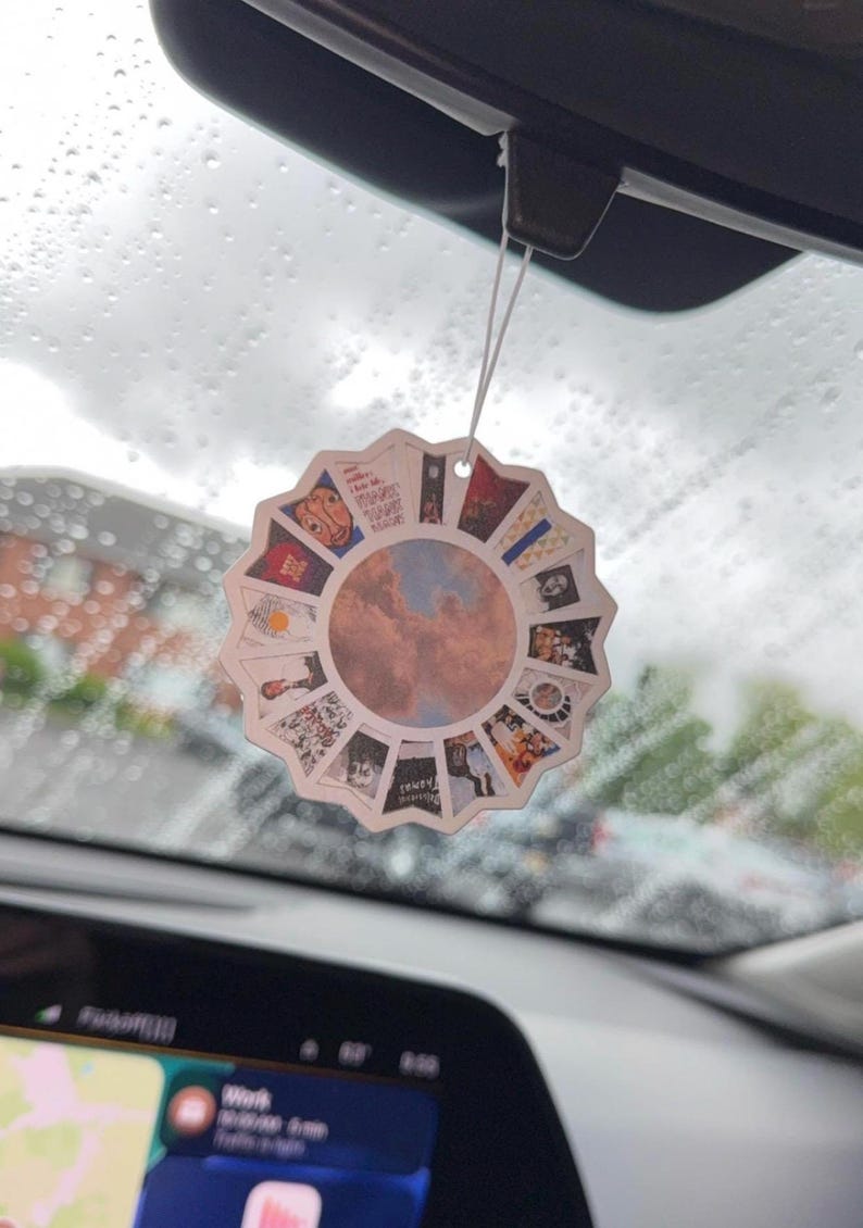 May include: A circular car air freshener featuring a scalloped edge and a central image of a cloudy blue sky. The border is composed of a collage of various small square album cover artworks arranged in a radial pattern around the centre.