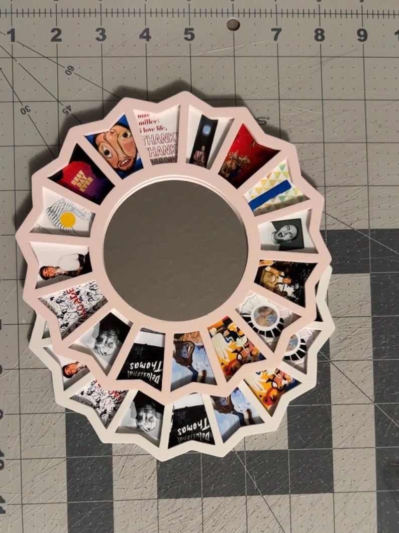 May include: A circular wall mirror with a light pink frame featuring a starburst edge. The frame contains multiple small, triangular photo inserts displaying various album art and imagery, including text reading Mac Miller and Delusional Thomas.