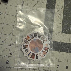 May include: A circular sticker featuring a central image of a cloudy sky surrounded by a ring of smaller, varied rectangular photographic prints, all enclosed within a clear plastic bag resting on a cutting mat with visible centimetre markings.