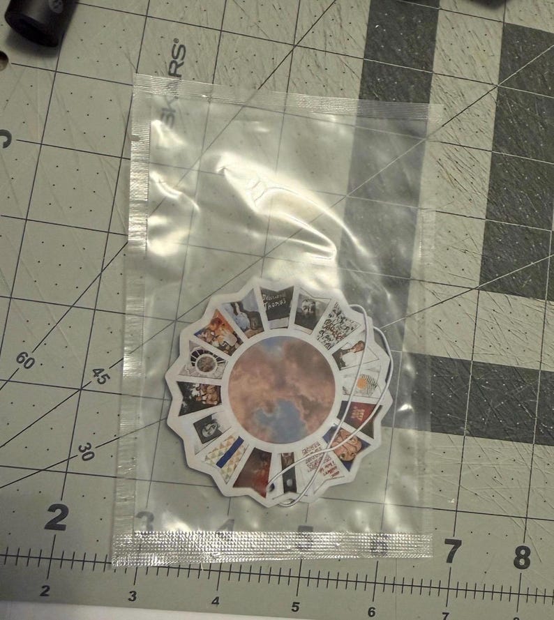 May include: A circular sticker featuring a central image of a cloudy sky surrounded by a ring of smaller, varied rectangular photographic prints, all enclosed within a clear plastic bag resting on a cutting mat with visible centimetre markings.