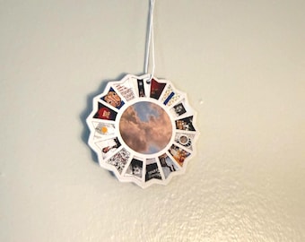 May include: A circular hanging ornament featuring a central image of a cloudy sky surrounded by a ring of sixteen small, distinct rectangular photo panels. The white-edged disc is suspended by a thin white string attached to the top centre.