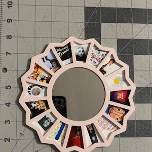 May include: A circular light pink frame with a scalloped edge surrounding a central mirror. The frame features twelve trapezoidal cutouts, each displaying a different small square image, including album art and portraits, arranged in a radial pattern.