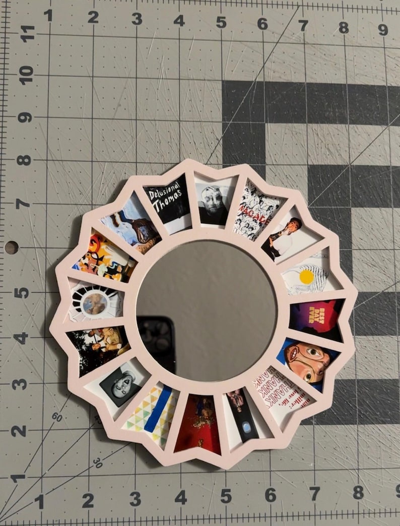 May include: A circular light pink frame with a scalloped edge surrounding a central mirror. The frame features twelve trapezoidal cutouts, each displaying a different small square image, including album art and portraits, arranged in a radial pattern.