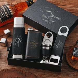 Puede incluir: Un juego de regalo negro para un padrino, que incluye una petaca, un abridor de botellas, un cortador de puros y un vaso de acero inoxidable. El juego está personalizado con el nombre "Bryan" y la fecha "01.09.25". También hay una botella de whisky y cubitos de hielo.