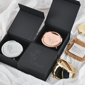 Puede incluir: Dos cajas de regalo negras abiertas con espejos compactos en su interior. Un espejo es plateado con un diseño floral y el nombre "Georgia". El otro es de oro rosa con el nombre "Daisy". También se ve un espejo compacto dorado y una pequeña etiqueta de arpillera.