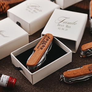 Może przedstawiać: Kolekcja spersonalizowanych prezentów dla drużbów, w tym drewniane multi-narzędzia i otwieracze do wina. Multi-narzędzia są grawerowane z imionami i słowem "Groomsman". Prezenty są prezentowane w białych pudełkach.
