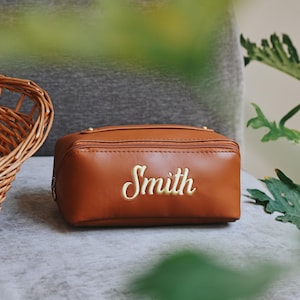May include: A brown leather cosmetic bag with the name "Smith" embroidered in gold script. The bag has a rectangular shape with a zipper closure. A wicker basket is visible in the background.