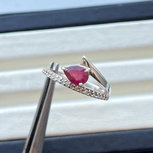 Könnte beinhalten: Ein silberner Ring mit einem birnenförmigen, tiefroten Edelstein. Das Band ist mit kleinen, klaren Steinen besetzt und hat ein einzigartiges, asymmetrisches Design. Der Ring wird von einer Pinzette gehalten.