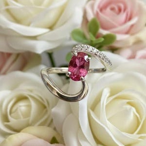 Könnte beinhalten: Ein silberner Ring mit einem großen, ovalen, rosa Edelstein. Der Ring hat ein geschwungenes Design mit einem Band aus kleinen, klaren Steinen. Der Ring ist vor einem Hintergrund aus weißen und rosa Rosen platziert.