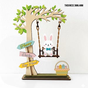 May include: Wooden Easter decoration featuring a tree with green leaves, a bunny on a swing, and a basket of eggs. The tree has three directional signs that read "Happy Easter Egg Hunt". The bunny wears a blue bow tie. The thickness is 3mm, 4mm.