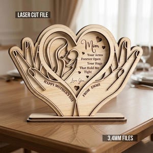 Könnte beinhalten: Eine lasergeschnittene Holzdekoration zum Muttertag. Das Design zeigt einen herzförmigen Ausschnitt mit Mutter und Kind, gehalten von Händen. Der Text enthält "Mom", "Happy Mother's Day" und "Love You". Die Basis ist 3,4 mm dick.