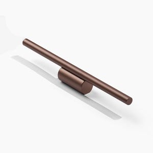 May include: A bronze-colored, minimalist wall clock with a cylindrical base and a long, straight bar forming the hour and minute hands. The clock is set against a white background, casting a shadow.