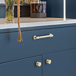 May include: Close-up of a navy blue cabinet with gold hardware. The cabinet features a long, horizontal handle and two smaller, round knobs. The hardware has a brushed gold finish, contrasting with the dark blue of the cabinet doors. The cabinet is part of a kitchen or bathroom setup.