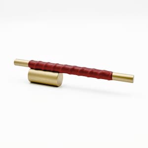 May include: A horizontal cabinet pull featuring a central cylindrical brass base and a long, straight handle. The handle has smooth brass ends and a textured, deep red leather-wrapped middle section that creates a distinct tactile grip.