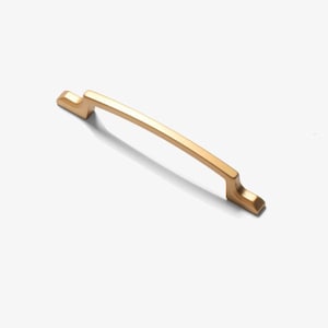 May include: A gold-colored cabinet pull with a curved bar and rectangular ends. The hardware is designed for easy gripping and is suitable for drawers or cabinet doors. The pull is made of metal and has a matte finish.