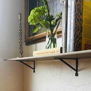 May include: A shelf with black metal brackets holds books, a vase of flowers, and a decorative candlestick. The books include "African Textiles" and "Behind the Blue Door". The shelf is mounted on a textured wall.