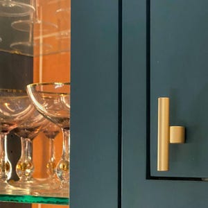 May include: A close-up of a teal cabinet door with a gold-colored handle. The handle is a vertical bar with a small, square knob. In the background, there are several champagne glasses with gold rims.