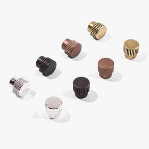 May include: An assortment of cabinet knobs in various metallic finishes, including silver, brass, and bronze. The knobs have a cylindrical shape with ribbed or smooth surfaces. The hardware is arranged on a white background.