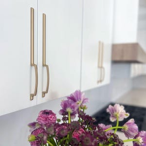 May include: Close-up of white kitchen cabinets with gold-toned handles. The handles have a unique, curved design. A bouquet of purple and pink flowers sits in front of the cabinets, adding a pop of color to the neutral kitchen setting.