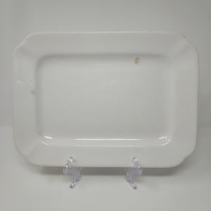 May include: A rectangular white ceramic serving platter with slightly scalloped edges. The platter is displayed on a clear plastic stand, showcasing its clean lines and simple design. A small, faint stain is visible on the surface.