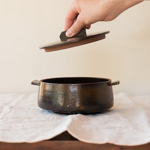 May include: A dark brown ceramic cooking pot with a matching lid. The pot has a textured surface and two small handles. The lid is being lifted, revealing the interior. The pot is sitting on a white cloth.