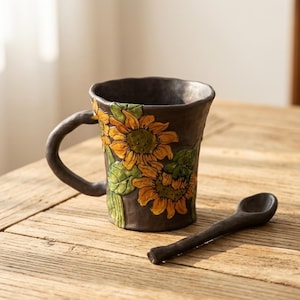 Puede incluir: Una taza de cerámica marrón oscuro con un diseño de girasoles y una cuchara a juego. Los girasoles son amarillos y naranjas con hojas verdes. La taza tiene una superficie texturizada y un asa curva. La cuchara es pequeña y tiene un cuenco redondeado.