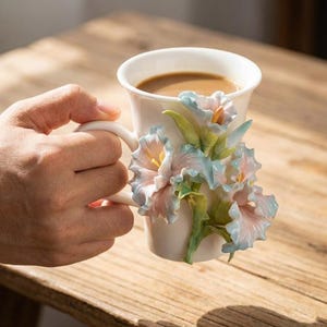 Könnte beinhalten: Weiße Keramiktasse mit Henkel, gefüllt mit Kaffee. Die Tasse ist mit 3D-Blumen in Rosa, Blau und Gelb verziert. Die Tasse wird von einer Hand gehalten und ruht auf einer Holzoberfläche.