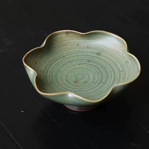 Op de afbeelding: Een handgemaakte, groene keramische kom met een unieke, geschulpte rand. De kom heeft een spiraalvormig ontwerp in het midden en kleine bruine spikkels. Een decoratief stuk voor in huis.