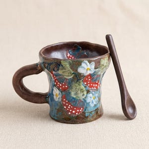 May include: A ceramic mug with a brown handle and a matching spoon. The mug is decorated with red strawberries, white flowers, and green leaves against a blue background. The mug and spoon are placed on a beige surface.
