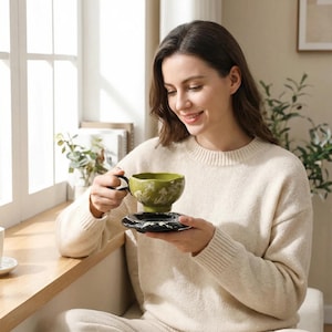 May include: A green ceramic teacup with a black handle and a matching saucer. The teacup features a floral design and is held by a person wearing a cream-coloured jumper. The teacup and saucer are on a wooden surface near a window.