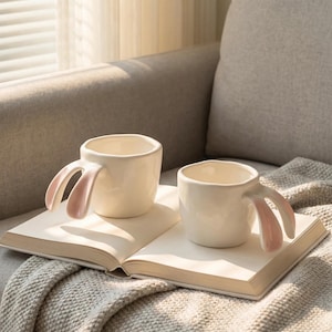 Puede incluir: Dos tazas de cerámica blanca con asas en forma de orejas de conejo rosas. Las tazas están sobre un libro abierto, sobre una manta suave, con un sofá al fondo.