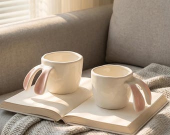 Peut inclure: Deux tasses en céramique blanche avec des anses en forme d'oreilles de lapin roses. Elles sont posées sur un livre ouvert, sur une couverture douce, avec un canapé en arrière-plan.