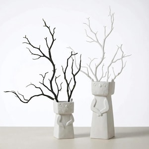 May include: Two white ceramic vases with cat-like faces and arms, each holding a bundle of branches. One vase holds black branches, the other white. The vases are decorative and suitable for home decor.