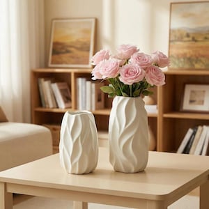 Puede incluir: Dos jarrones de cerámica blanquecina con un diseño texturizado ondulado. Un jarrón contiene un ramo de rosas rosas. Los jarrones están sobre una mesa de madera clara, con una estantería y obras de arte al fondo. Decoración del hogar.