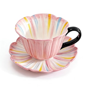 Puede incluir: Juego de taza y platillo de té de cerámica rosa con forma de flor. La taza y el platillo presentan rayas rosas, blancas, amarillas y azules. La taza tiene un asa negra. El juego está sobre un fondo blanco.