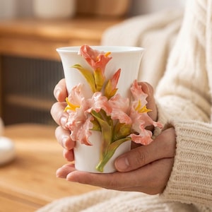 May include: White ceramic mug with a sculpted floral design in shades of pink, yellow, and green. The flowers are intricately detailed and appear three-dimensional. The mug is held in a person's hands.