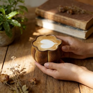 May include: A brown ceramic coffee cup with a flower-shaped rim, filled with a latte featuring a heart-shaped design. The cup is held in both hands, set against a wooden table with a plant and books in the background.