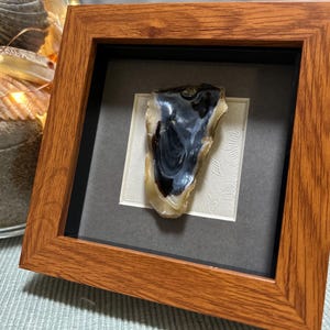 Puede incluir: Una piedra pulida enmarcada, con un llamativo patrón en blanco y negro. La piedra está montada sobre un fondo texturizado blanquecino dentro de un passe-partout gris, encerrada en un marco de madera cuadrado. El marco tiene un acabado marrón rojizo cálido.