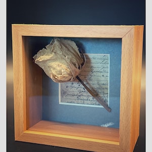 May include: A framed artwork featuring a dried rose with a brown stem, set against a vintage document. The rose is a light brown color, and the frame is made of light-colored wood. The document has handwritten text.