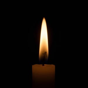 Könnte beinhalten: Eine brennende Kerze vor schwarzem Hintergrund. Die Flamme ist hellgelb-orange, mit einem etwas dunkleren Sockel. Die Kerze selbst ist cremeweiß.