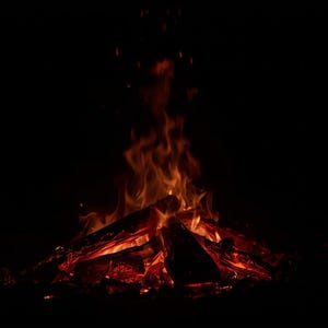 May include: A bonfire with bright orange and red flames against a black background. The fire is burning brightly, with sparks rising into the air. The wood is arranged in a conical shape.