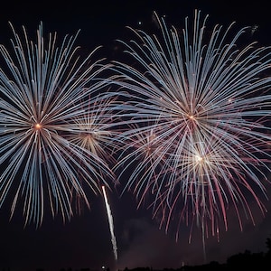 Puede incluir: Fuegos artificiales nocturnos. Dos grandes explosiones de fuegos artificiales azules y blancos dominan la imagen, con explosiones más pequeñas rojas y doradas. Un solo cohete asciende desde abajo.
