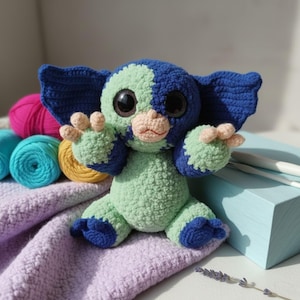 Op de afbeelding: Een handgemaakt gehaakt wezen in tinten blauw en groen, met grote zwarte ogen en kleine, bruine handen en voeten. Het pluche speelgoed heeft grote oren en zit op een paarse deken. Kleurrijke bollen garen zijn zichtbaar op de achtergrond.