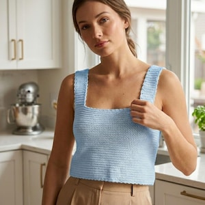 May include: A light blue crochet top with a square neckline and wide straps. The top is made of a textured yarn and has a cropped length. The model is wearing the top in a kitchen setting.
