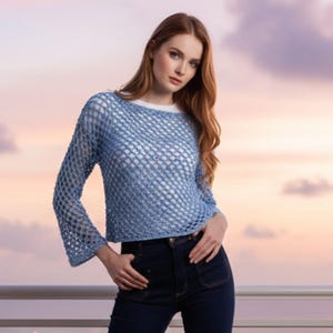 May include: A light blue, open-weave top with long sleeves. The top is paired with dark blue jeans. The top has a round neckline and a loose, airy design. The sleeves flare slightly at the wrists, creating a relaxed silhouette.